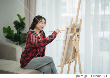 Young woman painting on canvas indoors, wearing casual outfit, focused on artwork, creativity at home, cozy atmosphere in living room Young woman painting on canvas indoors, wearing casual outfit, focused on artwork, creativity at home, cozy atmosphere in living room 136984971