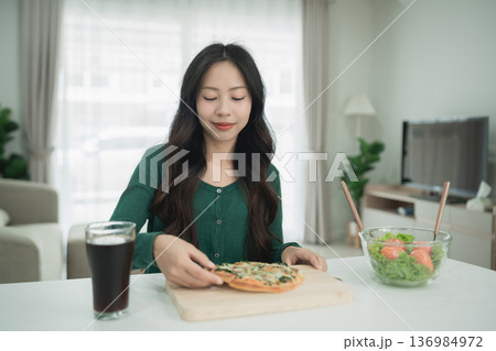 Young woman enjoying a homemade pizza while sitting at a bright kitchen table with a glass of soda and a bowl of fresh salad on a sunny day 136984972