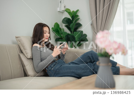 Young woman relaxing on couch while using smartphone in cozy living room with indoor plants and soft natural sunlight filtering through curtains 136986425