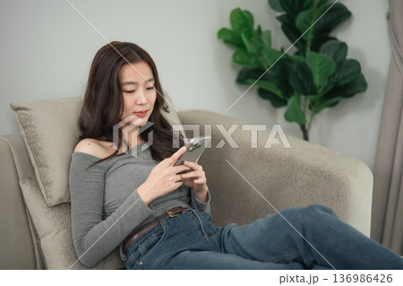 Young woman using smartphone while relaxing on couch in modern living room with indoor plants and cozy atmosphere for lifestyle or technology concept Young woman using smartphone while relaxing on couch in modern living room with indoor plants and cozy atmosphere for lifestyle or technology concept 136986426