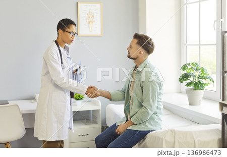 Clinic room meeting, female doctor shaking hands with male patient sitting on examination bed Clinic room meeting, female doctor shaking hands with male patient sitting on examination bed 136986473