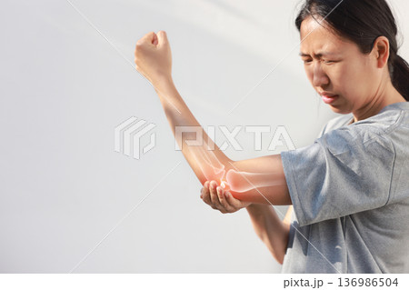 Asian woman suffering from elbow joint pain with red x-ray bone graphic overlay, female touching arm feeling ache from inflammation, arthritis, tendonitis or injury in garden, healthcare concept. 136986504