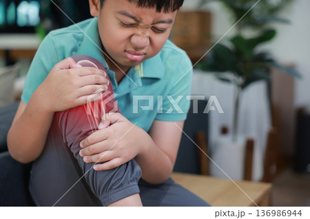 Little boy suffering from knee joint pain with red x-ray bone graphic overlay, child touching leg feeling ache from injury, growing pains, inflammation or accident while playing at home. 136986944