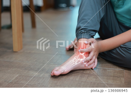 Little boy suffering from ankle joint pain with red x-ray bone graphic overlay, child touching foot feeling ache from injury, sprain, fracture or accidental fall while playing at home. 136986945