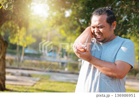 Adult Asian man suffering from elbow joint pain in park with red x-ray bone graphic overlay, male athlete touching arm feeling ache from tennis elbow, injury, inflammation or tendonitis. Adult Asian man suffering from elbow joint pain in park with red x-ray bone graphic overlay, male athlete touching arm feeling ache from tennis elbow, injury, inflammation or tendonitis. 136986951