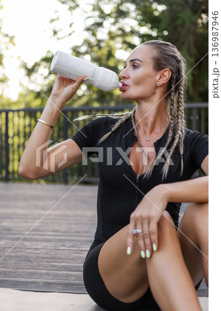 Athletic woman drinking water from sports bottle after outdoor workout outside in park. Fit female in black activewear resting in park, healthy lifestyle, hydration, fitness recovery 136987946