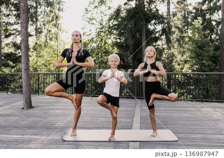Mother and children practicing yoga balance pose on mat outdoors in forest. Healthy family wellness activity with mindful exercise, calm focus and active lifestyle in summer. Mom with kids training Mother and children practicing yoga balance pose on mat outdoors in forest. Healthy family wellness activity with mindful exercise, calm focus and active lifestyle in summer. Mom with kids training 136987947