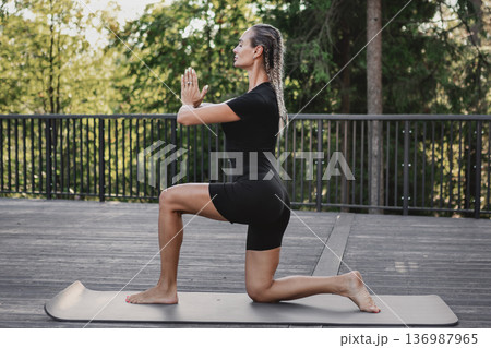 Athletic woman practicing low lunge yoga pose on mat outdoors in forest park. Female stretching hips and legs, mindful breathing, balance and flexibility training and wellness in nature 136987965