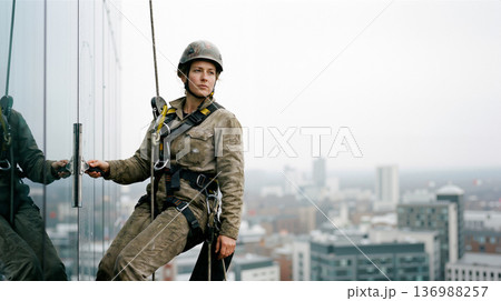 Professional Female Rope Access Worker Cleaning Glass on a Modern Building Skyscraper High Above the City 136988257
