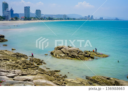 The landscape picture of the tranquil beach and azure sea at Hua Hin, Thailand with skyscraper view on the horizon line and blue sky. Beach rocks and tropical plants. 136988663