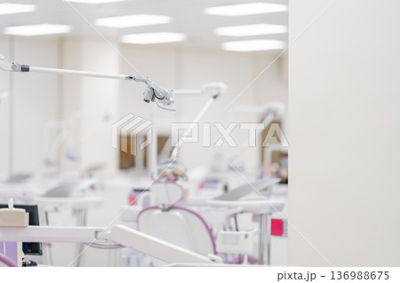 The side view of a modern white dental light lamp and reflector equipment above a medical chair on a blurry background of a dental clinic. The side view of a modern white dental light lamp and reflector equipment above a medical chair on a blurry background of a dental clinic. 136988675