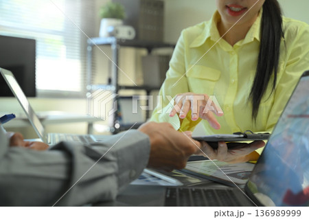 Close-up of coworkers discussing documents and digital data on a desk, showing teamwork, decision making, and professional collaboration in an office 136989999