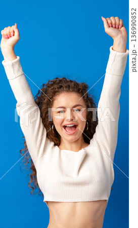 Cheering woman with curly hair raises arms and smiles against a blue background Cheering woman with curly hair raises arms and smiles against a blue background 136990852