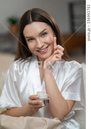 Young smiling woman applying hydrating cream on her face Young smiling woman applying hydrating cream on her face 136991781