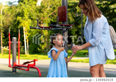 Mother helps daughter blow soap bubbles at playground park Mother helps daughter blow soap bubbles at playground park 136992019