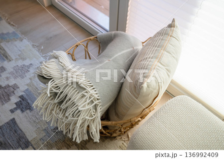 Rattan basket with soft blanket and pillow near window in cozy Scandinavian interior 136992409