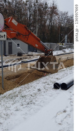 An orange excavator digs a dirt trench near black pipes in winter, construction site, industrial work. An orange excavator digs a dirt trench near black pipes in winter, construction site, industrial work. 136997669