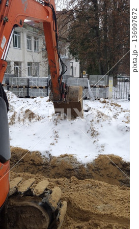 Close-up of an orange excavator bucket and tracks near a trench with pallets of paving slabs in the background, winter construction site, urban infrastructure. 136997672