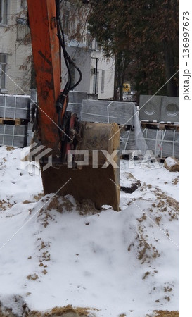 A close-up view of a dirty orange excavator bucket resting on a pile of snow and sand with building materials in the background, winter construction, heavy equipment. A close-up view of a dirty orange excavator bucket resting on a pile of snow and sand with building materials in the background, winter construction, heavy equipment. 136997673