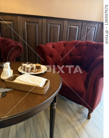 Small cakes and a napkin holder sit on a wooden table near red velvet armchairs, cozy cafe interior, relaxing lifestyle. 136997678