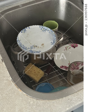 A dirty stainless steel kitchen sink is filled with unwashed pots plates and a sponge, messy home hygiene, daily household chores. 136997688