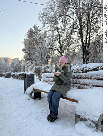A smiling woman sits on a snowy park bench holding a small snowball, winter leisure, outdoor lifestyle, cold season fun. A smiling woman sits on a snowy park bench holding a small snowball, winter leisure, outdoor lifestyle, cold season fun. 136997693