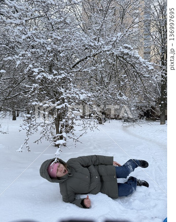 A woman lies happily in deep snow under a frosted tree, winter fun, playful outdoor lifestyle, frosty nature. 136997695