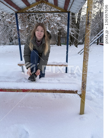 A woman sits on a snow-covered bench tying her shoelaces on a winter day, outdoor activity, cold season lifestyle, winter walk. 136997696