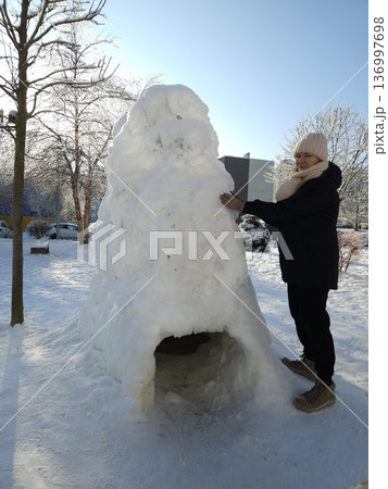 A woman stands next to a large snow fort with a tunnel entrance on a sunny winter day, outdoor recreation, creative winter fun, cold season activity. A woman stands next to a large snow fort with a tunnel entrance on a sunny winter day, outdoor recreation, creative winter fun, cold season activity. 136997698