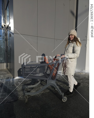 A woman pushes an empty shopping cart along a building on a sunny winter day, retail shopping, consumer lifestyle, outdoor errands. 136997704