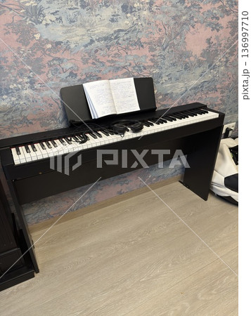 A black digital piano with open sheet music and headphones stands in a room with floral wallpaper, music education, creative hobby, home interior. A black digital piano with open sheet music and headphones stands in a room with floral wallpaper, music education, creative hobby, home interior. 136997710