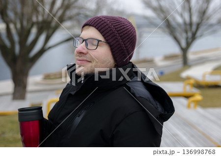 Young smiling man enjoying a warm drink from a red travel mug on a bank in winter. Concept of a city winter activity Young smiling man enjoying a warm drink from a red travel mug on a bank in winter. Concept of a city winter activity 136998768