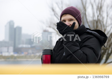 Young man enjoying a warm drink from a red travel mug near the river on a chilly day in winter. Concept of a city winter activity 136998769