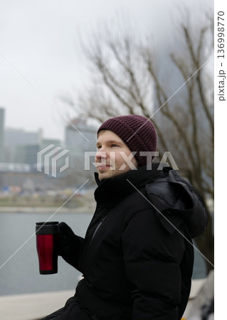 Young smiling man enjoying a warm drink from a red travel mug near the river on a chilly day in winter. Concept of a city winter activity. Young smiling man enjoying a warm drink from a red travel mug near the river on a chilly day in winter. Concept of a city winter activity. 136998770