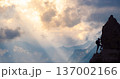 A climber scales a rocky mountain peak as the sun sets, casting a golden glow over the landscape filled with distant peaks and clouds. 137002166