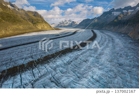 A vast glacier Aletsch Glacier in Switzerland Aps flows between rugged mountains on a bright summer day. The beautiful landscape showcases stunning natural formations and greenery. Aerial drone view. 137002167
