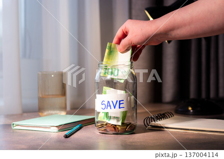 Close up of cash being added to transparent money jar. Represents traditional saving methods and conscious money management for future security 137004114