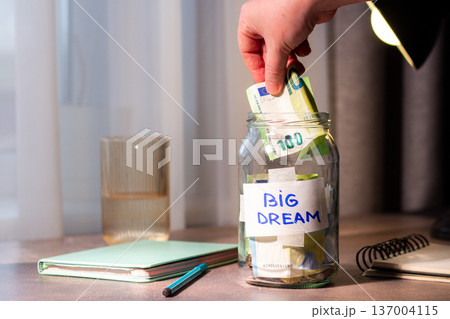 Hand dropping money into glass savings jar on table. Close up of fingers inserting cash into transparent container for big dream. Financial planning and goal achievement concept 137004115