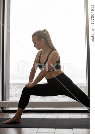 A fit woman in black athletic wear performs a deep lunge stretch on a yoga mat in front of a large window, showcasing flexibility and dedication to fitness 137004615