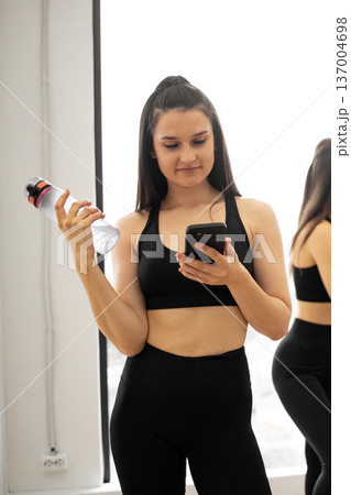 A fit woman in athletic wear holds a water bottle and checks her smartphone after a strenuous exercise session A fit woman in athletic wear holds a water bottle and checks her smartphone after a strenuous exercise session 137004698
