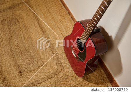 Red Acoustic Guitar Leaning Against Wall on Woven Rug in Warm Interior Red Acoustic Guitar Leaning Against Wall on Woven Rug in Warm Interior 137008759