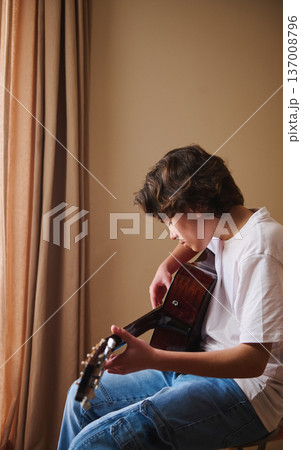 Teen Boy Playing Acoustic Guitar at Home by a Window, Practicing Music and Concentration Teen Boy Playing Acoustic Guitar at Home by a Window, Practicing Music and Concentration 137008796