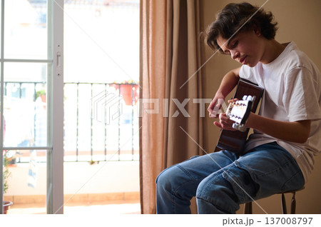 Teen Boy Playing Acoustic Guitar Indoors Near Balcony Window During Casual Home Practice Teen Boy Playing Acoustic Guitar Indoors Near Balcony Window During Casual Home Practice 137008797