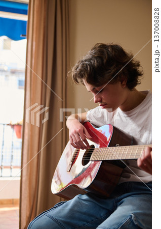 Teen Boy Playing Acoustic Guitar Indoors Near Window Practicing Music at Home Teen Boy Playing Acoustic Guitar Indoors Near Window Practicing Music at Home 137008828