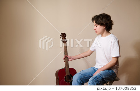 Teen Boy Sitting With Acoustic Guitar Practicing Music in Casual White T-Shirt and Jeans 137008852