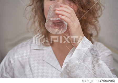 Young caucasian female drinking water in white pajamas at home for morning hydration routine 137008968