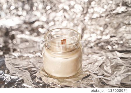 Small glass candle with wooden wick on reflective foil creating cozy ambient light in soft focus Small glass candle with wooden wick on reflective foil creating cozy ambient light in soft focus 137008974