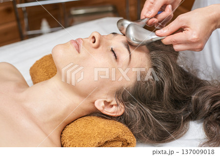 Young Caucasian Female Receiving Relaxing Facial Treatment With Metal Spa Tools For Rejuvenation 137009018