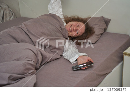 Caucasian female adult waking up smiling in bed holding smartphone with clock showing 5:00 137009373