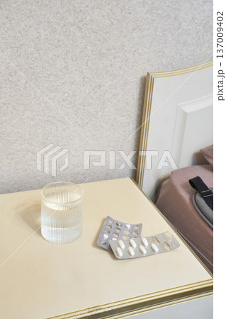 Glass of water and empty medicine blisters on nightstand beside bed with beige headboard in bedroom 137009402